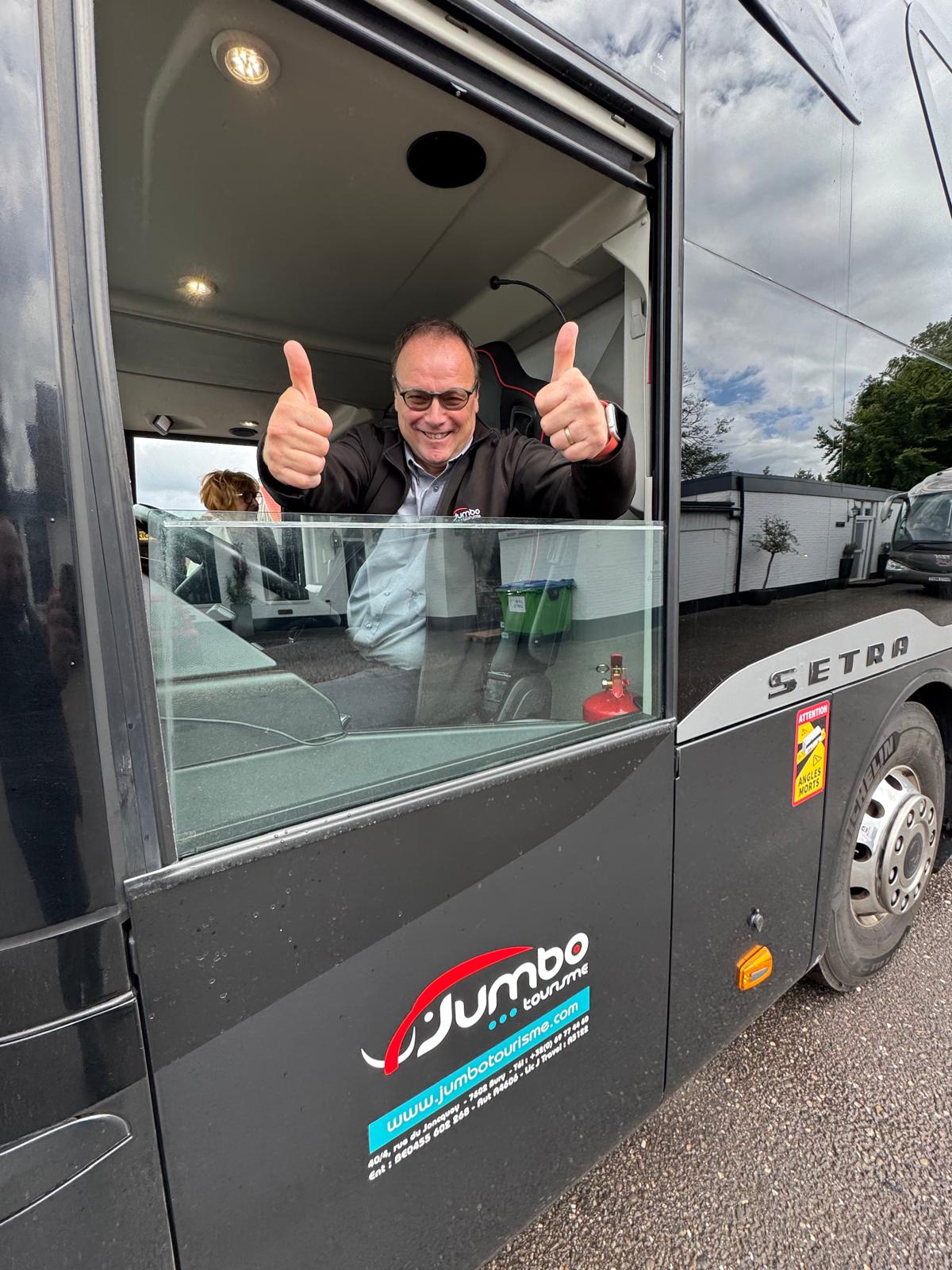 Chauffeur Jumbo Tourisme à la fenêtre de son autocar, pouces levés.